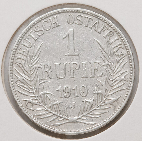 Die Rückseite der Münze zeigt den Nennwert „1 RUPIE“ und das Prägejahr „1910“ im Zentrum. Diese Angaben werden von einem Kranz aus zwei unten zusammengebundenen Palmzweigen umschlossen. Am oberen Rand steht die Inschrift „DEUTSCH OSTAFRIKA“ und unter der Jahreszahl ist das Münzzeichen „J“ für Hamburg zu sehen.