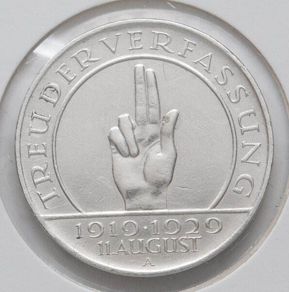Nahaufnahme der Motivseite der 3-Reichsmark-Gedenkmünze von 1929. Gezeigt wird die Schwurhand mit drei gestreckten Fingern im Zentrum. Die Umschrift lautet TREUE DER VERFASSUNG und unten das Datum 11. AUGUST. Unter der Jahreszahl 1919-1929 ist das Münzzeichen A für Berlin sichtbar.