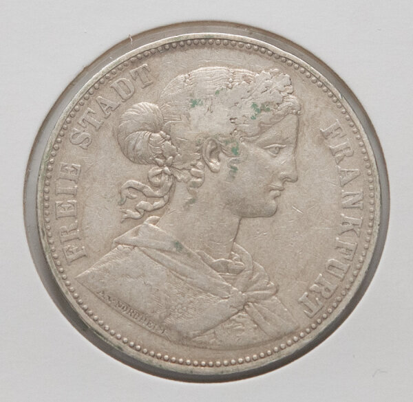 Nahaufnahme der Vorderseite des Frankfurter Vereinstalers von 1865. Abgebildet ist das Brustbild der Francofurtia mit Mauerkrone und kunstvoller Frisur. Die Umschrift lautet FREIE STADT FRANKFURT. Am Halsabschnitt ist die Signatur des Künstlers Nordheim erkennbar.