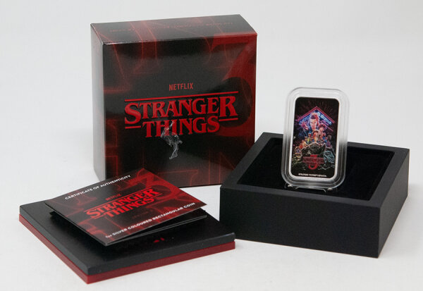 Kompletter Lieferumfang: Der colorierte Silberbarren in der Schutzkapsel, gebettet in einem schwarzen Display-Halter, daneben das rote Zertifikat und die thematisch gestaltete Stranger Things Umverpackung.